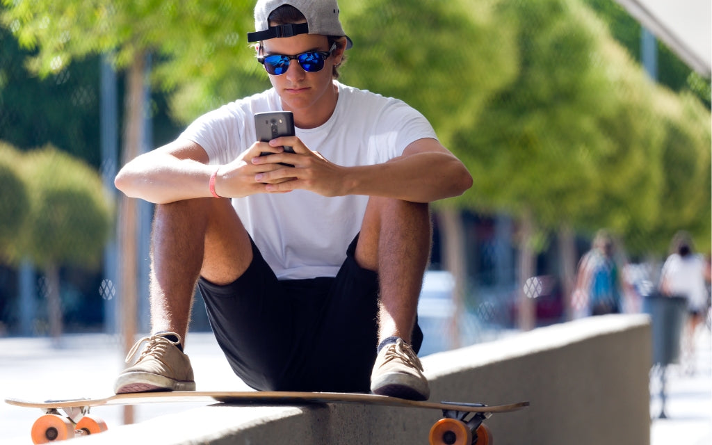 Youthful Guy in Blue Sunglasses Resting on Concrete Median With His Skateboard and Looking at His Smartphone