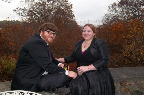 A brown haired man with a red beard, seated on a stone wall wearing a suit, next to a red haired woman in a fancy black dress, also seated on the stone wall. Both are smiling as if laughing.Fall foliage can be seen in the background.