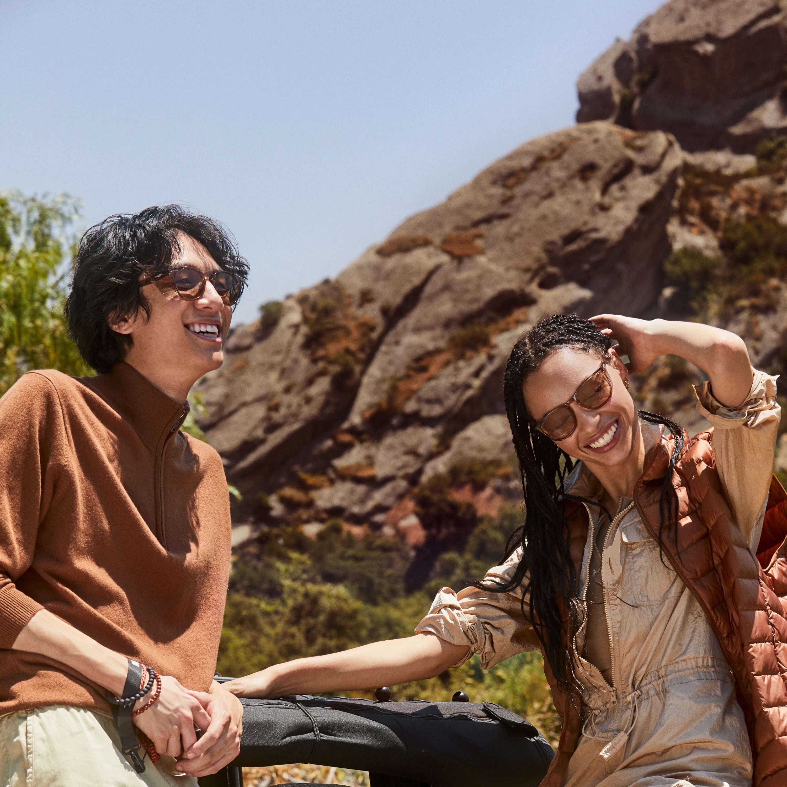 Two smiling people wearing SunStoggles sitting outdoors against large rock formations on a clear day.