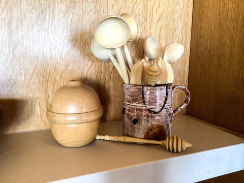 A small piece of pottery sits upon a shelf, holding 6 handmade spoons, 3 honey spinners from morrocco and two lemon juicers. Beside it sits a salt pig. 
