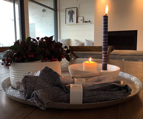Two candles are lit on a dining table, they are sitting in a porcelain bowl. Carefully placed between napkins, plants and a cup of coffee. 