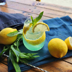 Gin Limoncello Collins with mint cocktail recipe by Newy Distillery. Gin Limoncello Collins cocktail in gin glass, with lemon and mint garnish, displayed on blue cloth with fresh mint, lemons and stirring spoon.