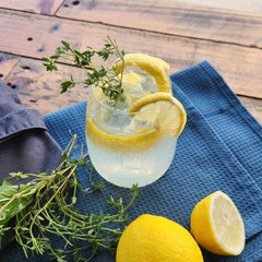 Gin Limoncello Collins with thyme cocktail recipe by Newy Distillery. Gin Limoncello Collins cocktail in gin glass, with lemon and thyme garnish, displayed on blue cloth with fresh thyme, lemons and stirring spoon.