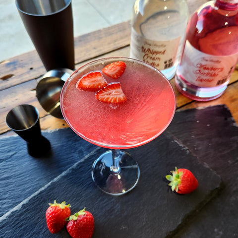 Strawberry Shortcake Martini Cocktail Recipe by Newy Distillery. Strawberry Shortcake Martini pink in colour, served in martini glass with sliced strawberry garnish. On a grey slate with strawberries, cocktail shaker and Newy Distillery Whipped Cream vodka bottle and Strawberry Shortcake Liqueur Vodka Bottle.