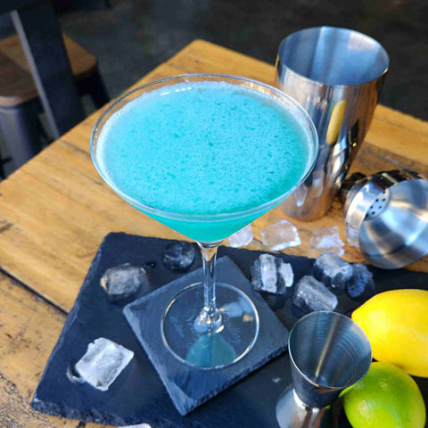 Aqua Vodka Martini Cocktail by Newy Distillery. Turquoise Blue cocktail in martini glass, on wooden table. Lime, lemon and cocktail shaker and ice surround. Cocktail recipes by Newy Distillery.