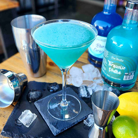 Aqua Vodka Martini by Newy Distillery. Blue vodka martini cocktail in martini glass on wooden table. Surrounded by lemon. lime, cocktail mixer, Newy Distillery Turquoise Vodka 700ml, Newy Distillery Blue Curacao 700ml.