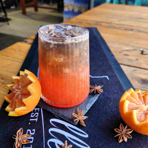 Newy Distillery Lava Cloud Cocktail. Vodka ocktail drink made with Newy Distillery Black Liquorice Vodka and Orange flavoured Vodka. Lava Cloud cocktail displayed on black bar mat on wooden table. Decorated with orange halves and star anise.