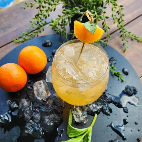 Mandarin Gin Collins Cocktail Recipe. Made with Newy Distillery Mandarin Gin. Mandarin Gin Collins cocktail with mandarin orange and mint garnish on a black chopping board, surrounded by ice, mandarin oranges and mint sprig.