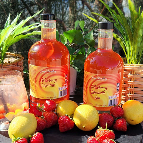 Newy Distillery Strawberry Limoncellos Double Release. Strawberry Limoncello Gin 500ml and Strawberry Limoncello Liquuer 700ml. Bottles side by side surrounded by lemons and strawberries. Green plants in the background.