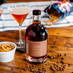 Newy Distillery Caramel Espresso Martini Pre-Mix Cocktail Bottle 700ml. Ready to serve Caramel Espresso Martini Bottle displayed on brown wooden table next to small pot of pretzels and scattering of coffee beans. Two Caramel Espresso Martini cocktails served. Bag of pretzels on white cloth in the background.