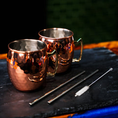two copper moscow mule mugs with straws and straw-cleaner presented on grey slate. Newy distillery. 