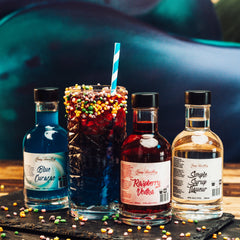 Newy Distillery Fruit Tingle Cocktial Kit. Make Fruit Tingle cocktails at home. Fruit Tingel cocktail in tall glass with straw and Nerds candy garnish. Served in grey slate with Nerd candt scattered around. 200ml Newy Distillery Blue Curacao on left. 200ml Newy Distilllery Raspberry Vodka and 200ml Newy Distillery Simple Syrup on the right.