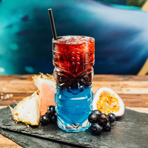 Tropical Punch cocktail. Blue and red cocktail served in tiki glass surrounded by tropical fruit. Newy Distillery cocktails.