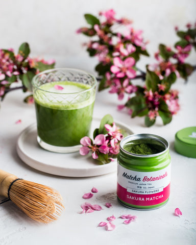 Sakura matcha in pink and green tin next to a glass of green matcha with pretty pink flowers surrounding