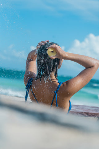 Hair Cooki lemon shampoo bar being used by the blue ocean on a sandy beach. Girl has brown hair and wears a blue bikini