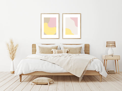 Set of Two Abstract Pastel Art Prints in Pinks, Yellows and Light Brown Neopolitan Colours in a Bedroom Setting with Bed, Bedside Table and Dried Grasses 