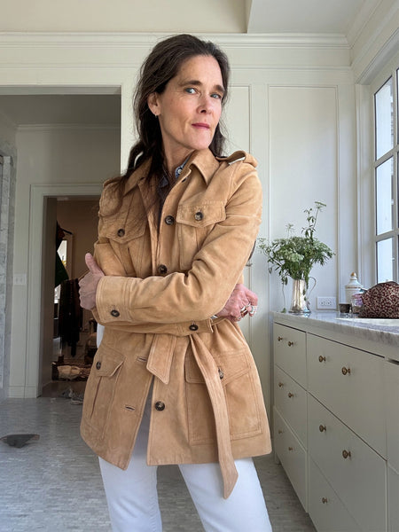 Ann at home in her Mashburn suede safari jacket. The jacket is made up in a rich camel-colored lamb suede with flap pockets, horn buttons, and a self-tie belt (very YSL.) In the background, there's a big bunch of spring flowers in a pitcher.