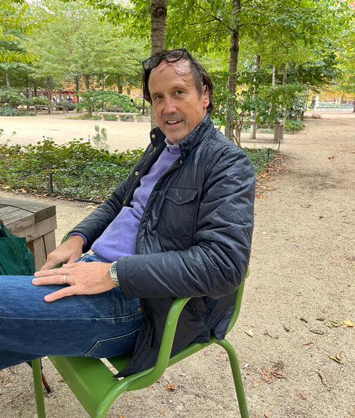 Sid sitting in a chair in the Tuilleries garden in Paris in what would make a pretty great tailgating outfit: a lightweight, navy cashpad jacket layered over a plaid sport shirt and a lavender fine-gauge sweater, paired with indigo rinse denim..