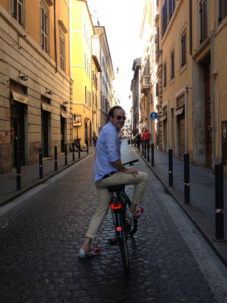 Sid biking down the streets of Rome in an OCBD, khaki trousers, and a pair of trainers.