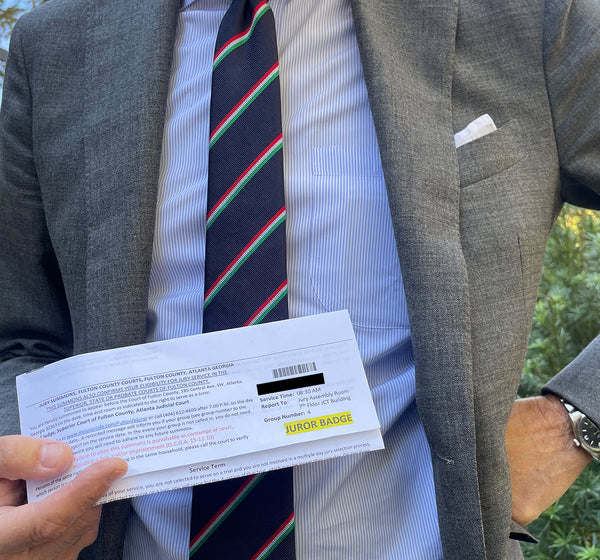 Sid in a grey wool suit, blue dress shirt, and silk repp tie holding his recent summon for jury duty. He has a white pocket square in his left suit pocket.