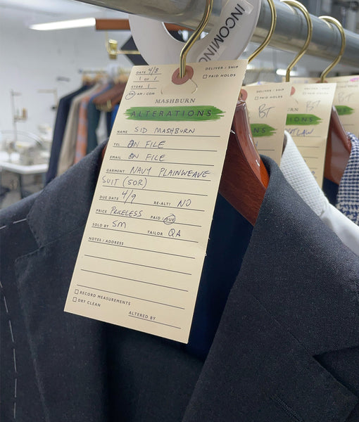 Sid's Navy Plainweave suit in the queue for alterations. It's tagged with handwritten specs, and in the background you can make out a sewing machine.