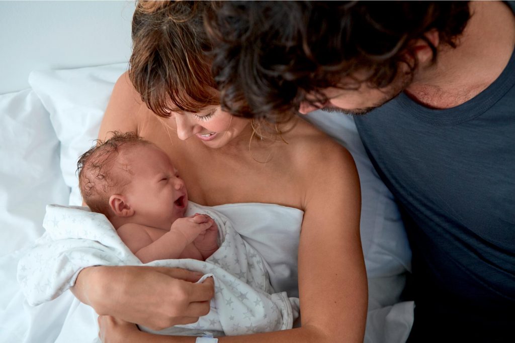 A mother sits in bed with her newly born baby. The father is stood next to them with his arm round the mum, smiling at them both.