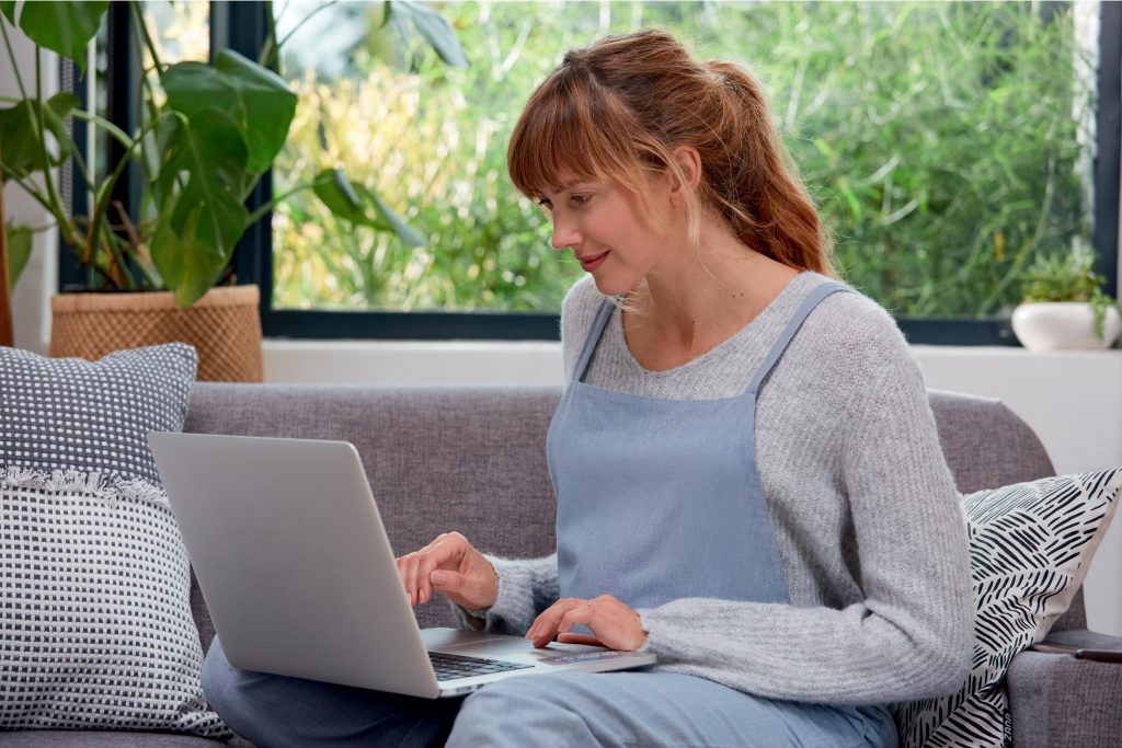 A pregnant woman is sat on a sofa in a brightly lit living room with an open laptop. She is typing on the laptop.