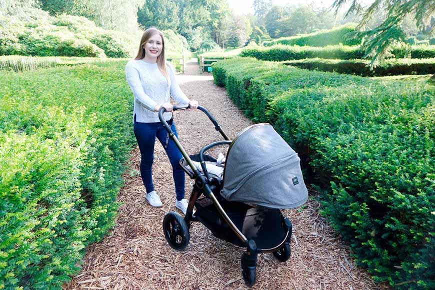 Haley is posing with the Ocarro Simply Luxe pushchair in a park, on a gravel path, between rows of bushes. 