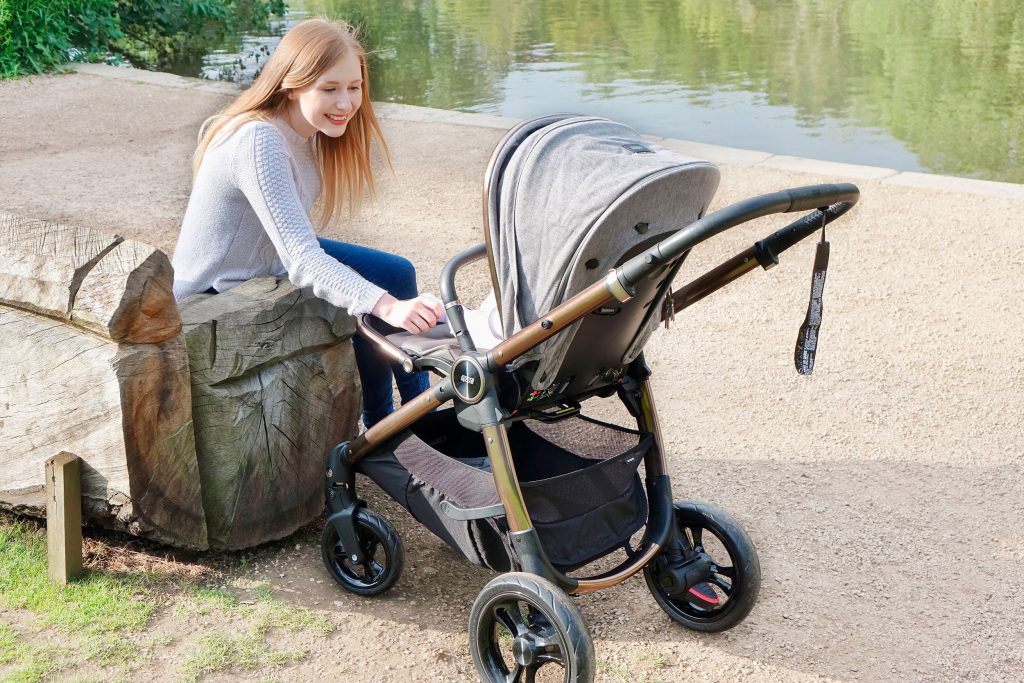 Haley is sat on a park bench next to a pond, she has the Ocarro pushchair next to her and she is entertaining baby Elodie who is sat inside.
