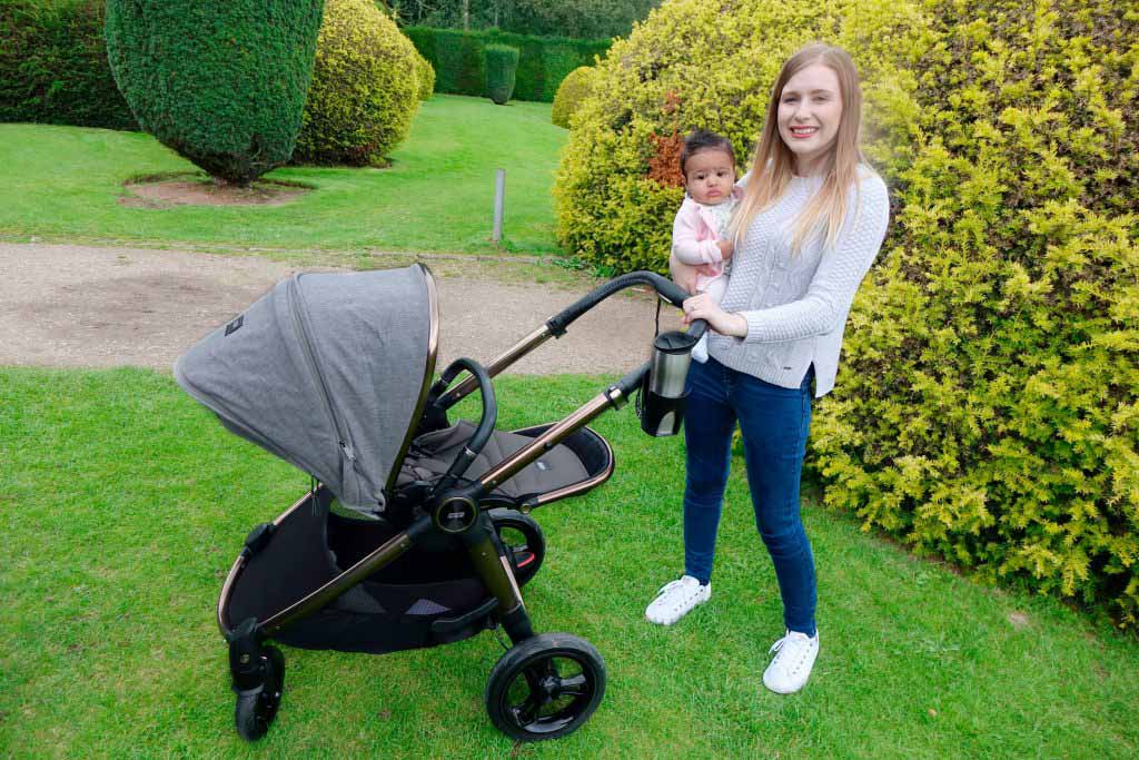 Haley is carrying baby Elodie and they are posing with the Ocarro pushchair, behind them is a hedge.