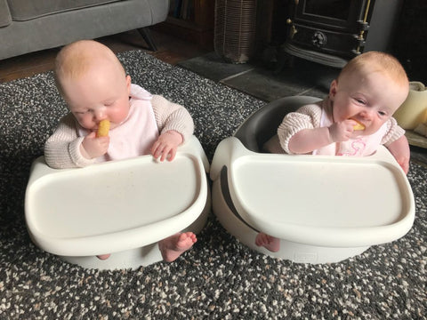 The young twin girls, Esther and Eve, are sat in their Baby Snug booster seats and are both chewing on a piece of food.
