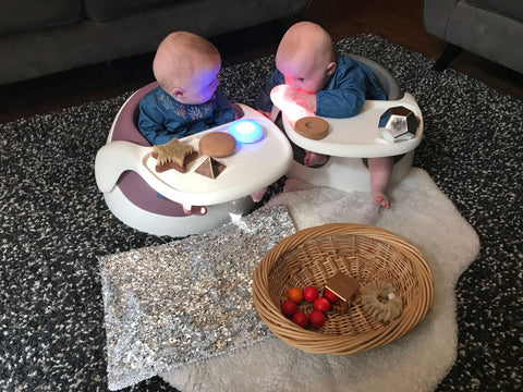 Eve and Esther are sat in their Baby Snug booster seats, surrounded by interactive toys and objects.