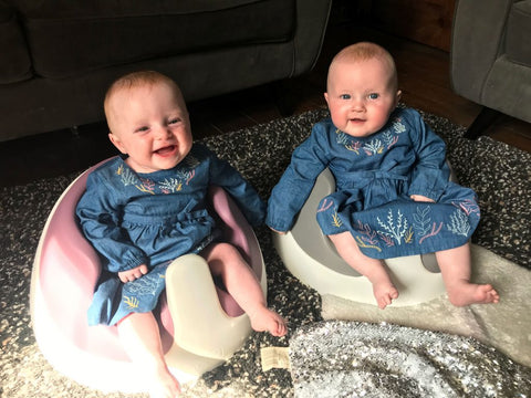 Twin girls, Esther and Eve are wearing matching denim dresses and are sat on the floor in their Baby Snug booster seats.
