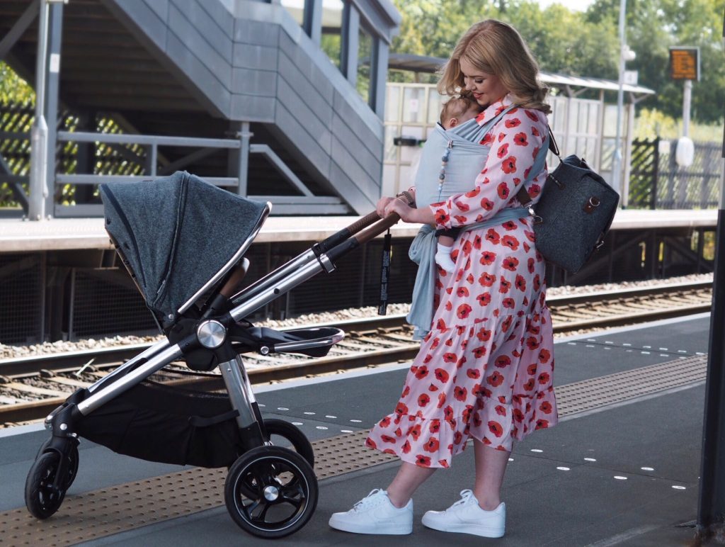 Image of Harley on a train station platform pushing the Ocarro Moon pushchair, while baby Freddie is fast asleep in a baby carrier strapped to Harley's chest.