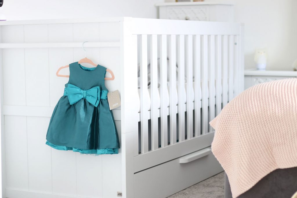 Image of a Franklin cot bed in a parent's bedroom, with a green occasionwear dress handing from the hanging rail along the side.