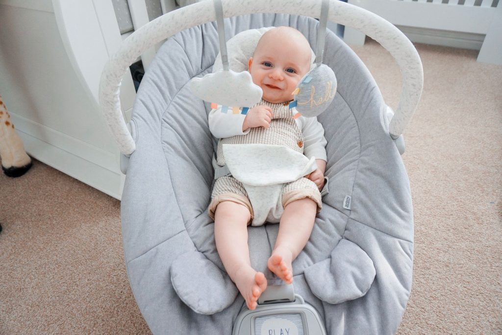 Baby Theo is sat in his Apollo cradle, in the middle of a nursery. The cradle has a large plush looking seat and a handle that reached over him. Attached to the handle are two soft hanging toys.