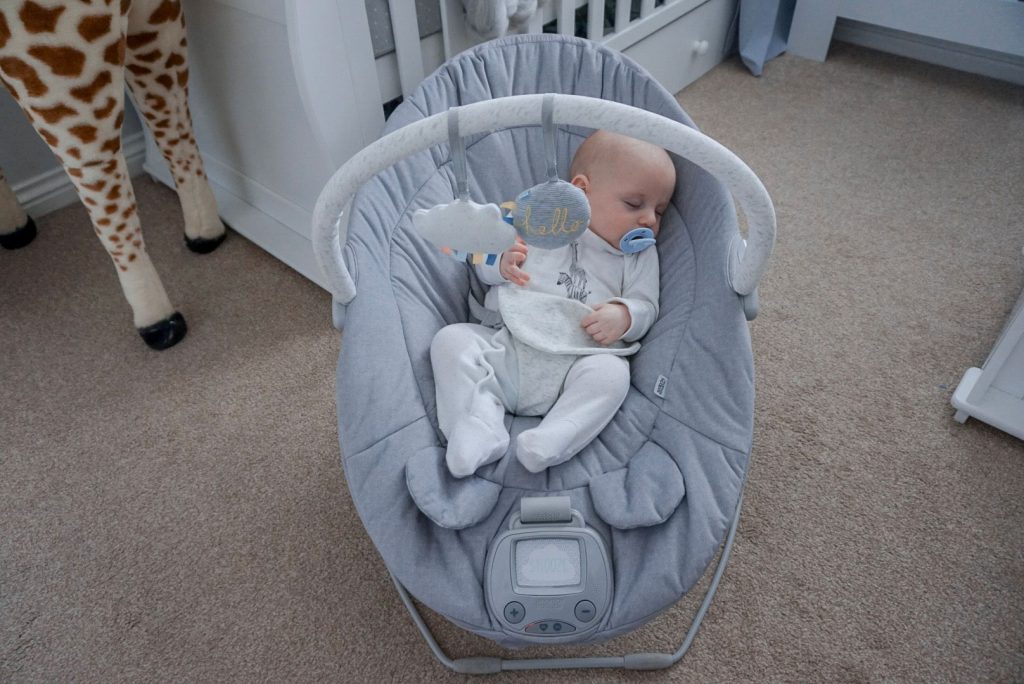 Another image of baby Theo asleep in the cradle, in the middle of a nursery. Behind thew cradle is a cot bed and dresser.
