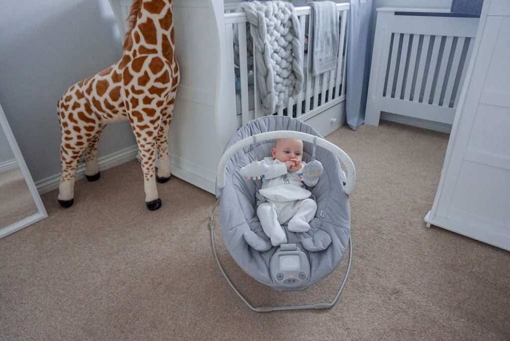 Baby Theo sat playing in his Apollo cradle in the middle of a nursery. Behind him is a cot, a mirror, a dresser changer and a large soft toy of a giraffe.