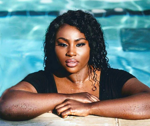 An African American woman is pictured with her arms crossed over the side of the pool with her shoulders up pictured only. Behind her the clear blue water of the pool is visible.