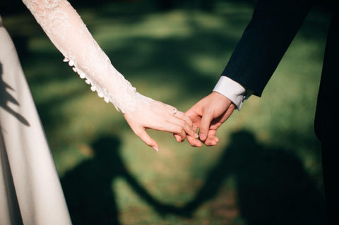 A picture of two people's arms reaching out for each other and connected only by the grasp of a few fingers. One arm is pictured in a suit jacket while the other person appears to be wearing a wedding dress. The background is green trees.