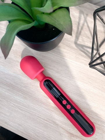 A bright pink rubber wand shaped object that is two thirds black is laying flat on a pale wood bench pictured from above and with a potted plant in view beside it.