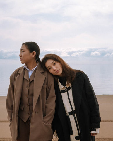 A female couple pictured, both wearing warm coats, and one has her head placed on her partners shoulder. Behind them in an image of the sky and a clear, calm, clear lake.