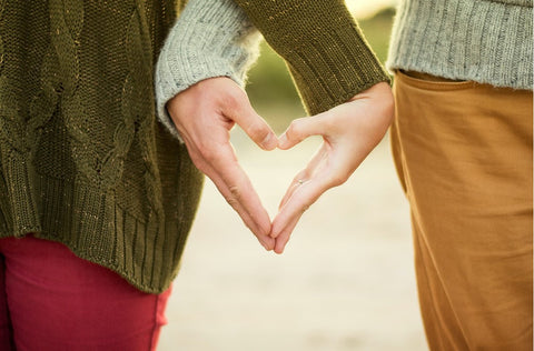 A couples hands are pictured close up with the sides of their bodies the only other thing visible and their hands are together in the shape of a heart.