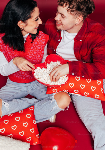 A couple are pictured on a red background and they are both wearing red. They are sitting on a couch looking into each others eyes and smiling while sharing a bowl of popcorn which is placed in between them and they each have a hand in the bowl.