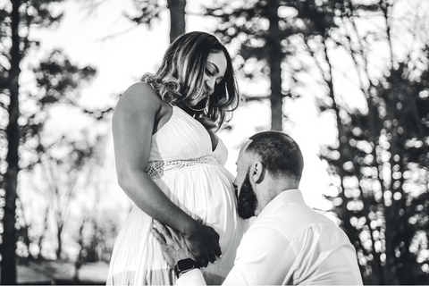 A black and white image of a couple on their wedding day pictured from the waist up. The man is kneeling at the woman's feet and is holding her hands while kissing her very pregnant stomach. Behind them in the background are tall trees.