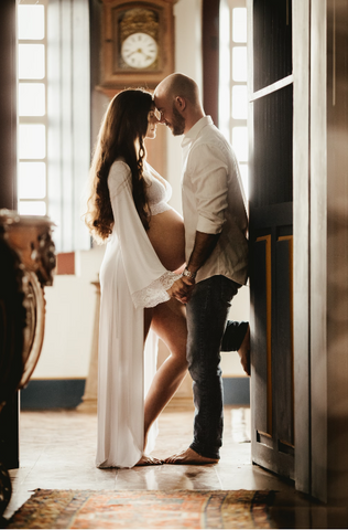 A couple are pictured standing, facing each other, holding hands and with their foreheads touching. The woman is wearing a long white dress with her big pregnant stomach sticking out while the man is wearing jeans and a white linen shirt.