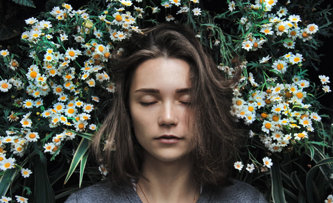 A woman with short brown hair is pictured from the shoulders up facing towards the camera but with her eyes closed. Behind her on either side of her head is a huge bunch of tiny white daisys.