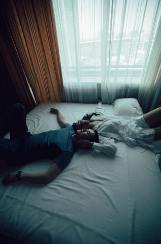 A couple are pictured in bed together looking up at the celiing with their heads touching but bodies facing opposite directions. The woman's hand is gently resting on the man's cheek while he has both his arms at his sides.