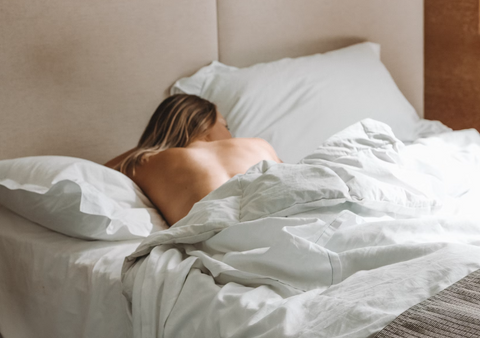 A women is laying in bed on her stomach and facing away from the camera. She is laying on white sheets with the white cover pulled up to her lower back with the rest of her bare back exposed.