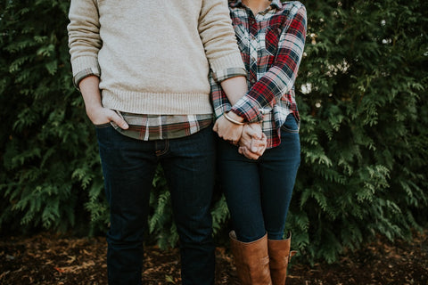 Couple facing forward but cut off at the shoulders and holding hands. Man is wearing a cream jumper while the woman is wearing a checked shirt and both in jeans.